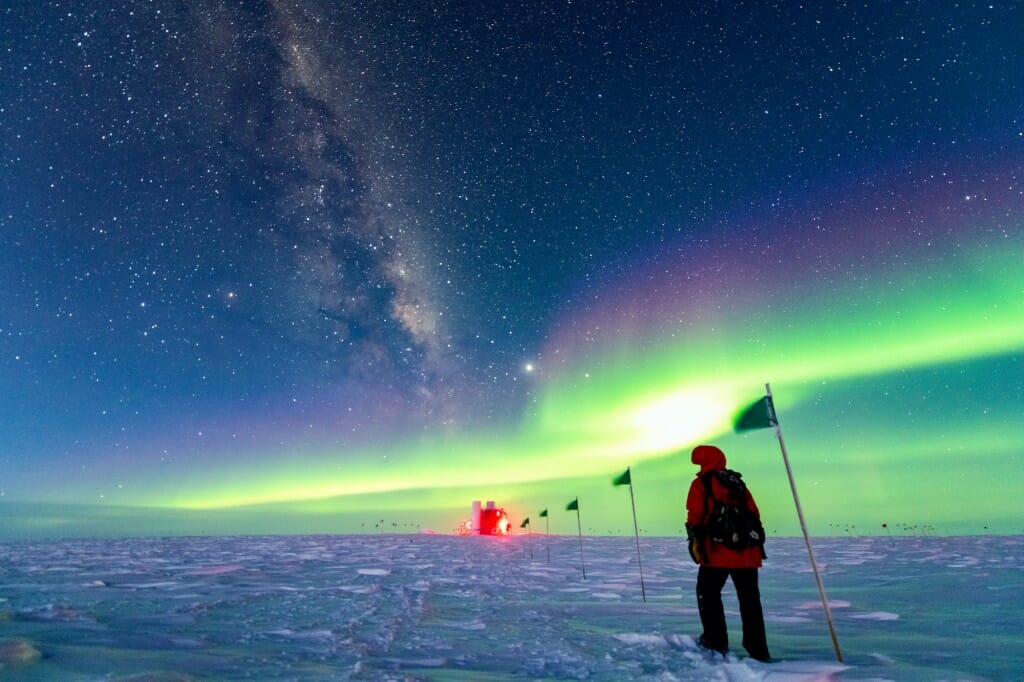 Person walking toward observatory over snow, under the Southern Lights.