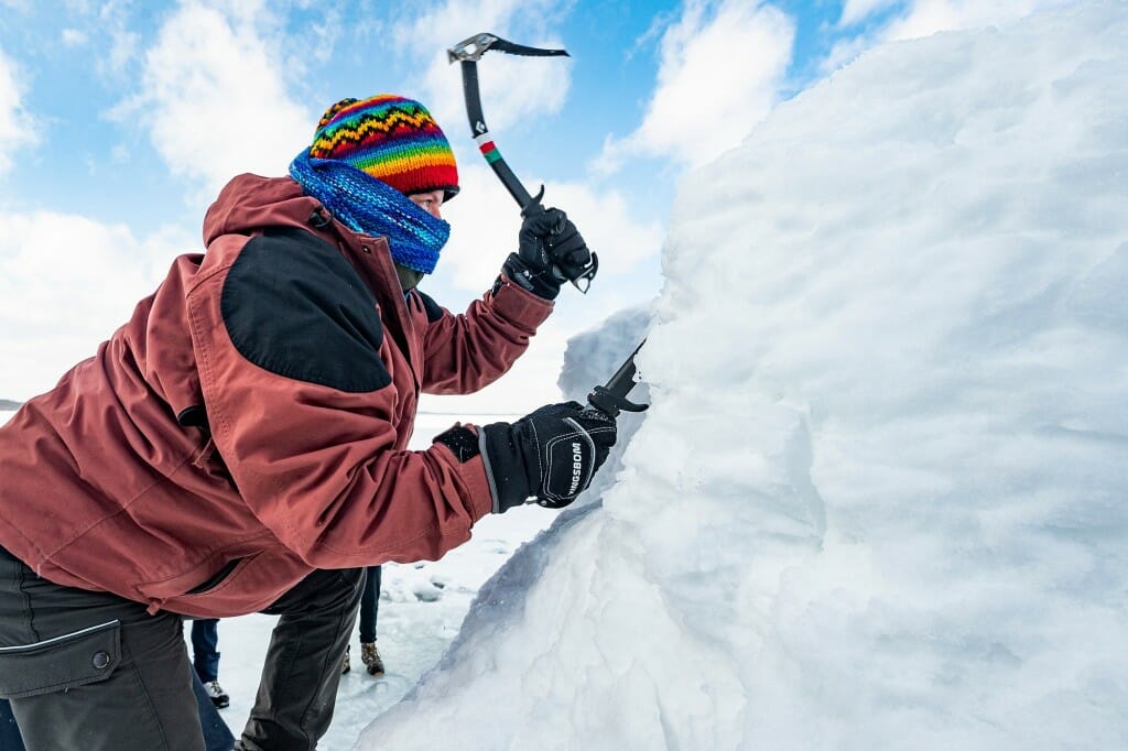 A person dressed in warm winter clothes pounding a pick into a pile of ice