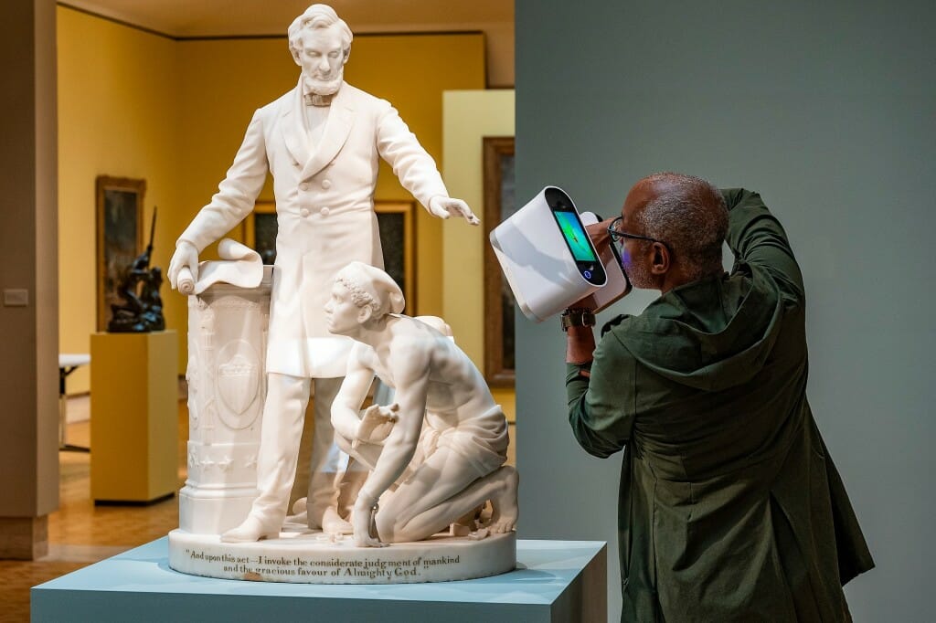 A person aiming a 3-D imaging device at a statue of Abraham Lincoln with an enslaved person crouching next to him