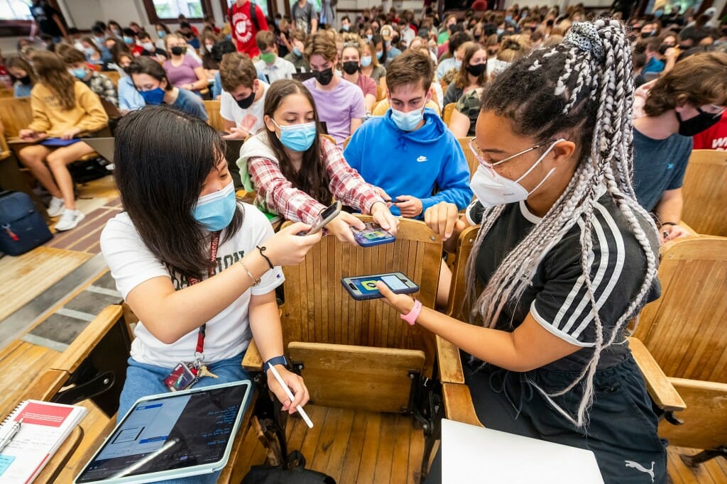 Several students in a large classroom reaching to each other with their phones