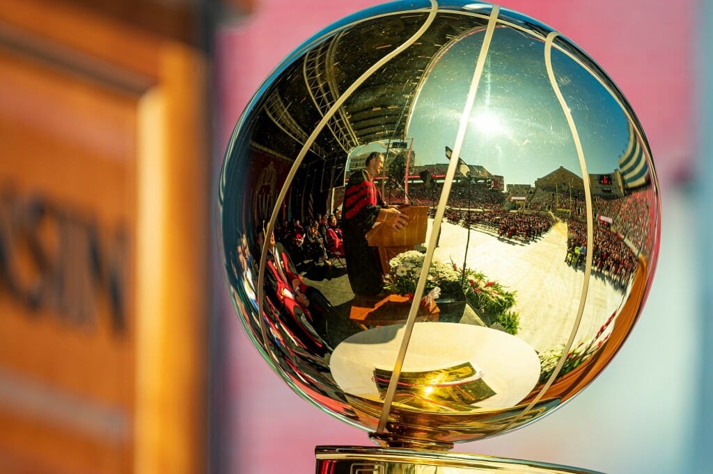 Closeup of basketball-shaped trophy with reflection of Connaughton, wearing academic gown and speaking at podium