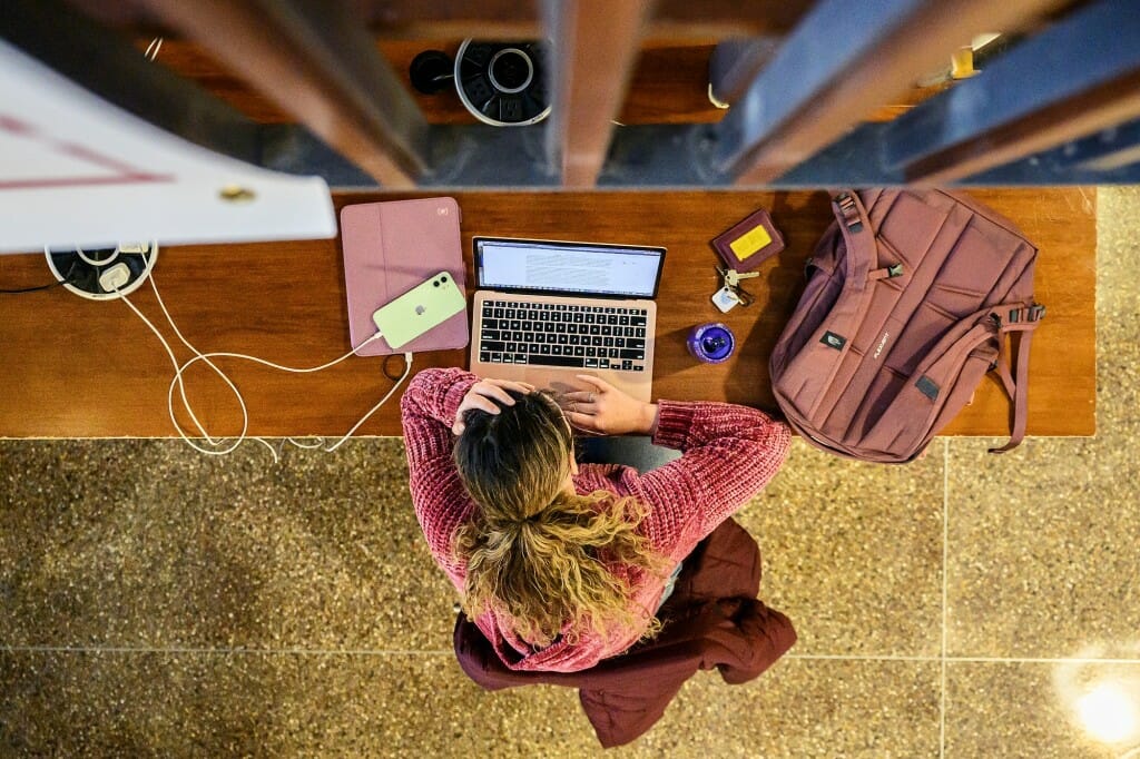 Overhead view of a student at a table with laptop open next to plugged-in tablet and phone, a backpack and a set of keys