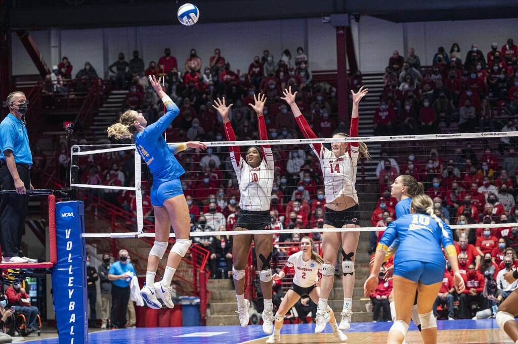 Two Wisconsin players raise their arms as a UCLA player prepares to hit the ball