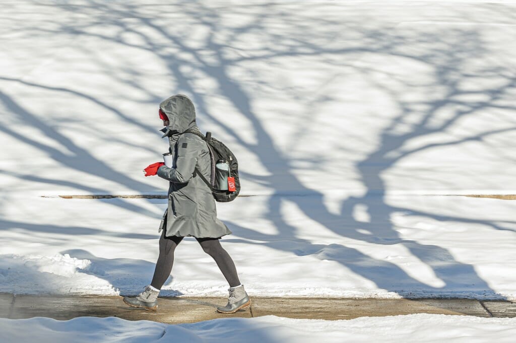 A student wearing a gray parka and bright red gloves walks next to a snowy lawn.