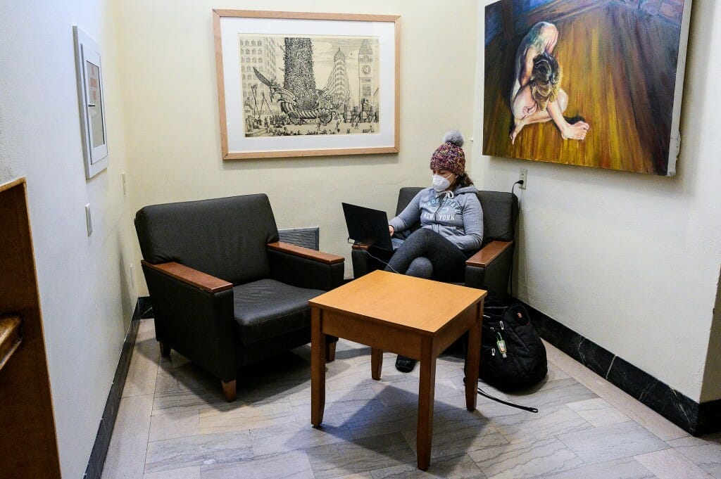 A student wearing a face mask sits in a cozy chair working on her laptop