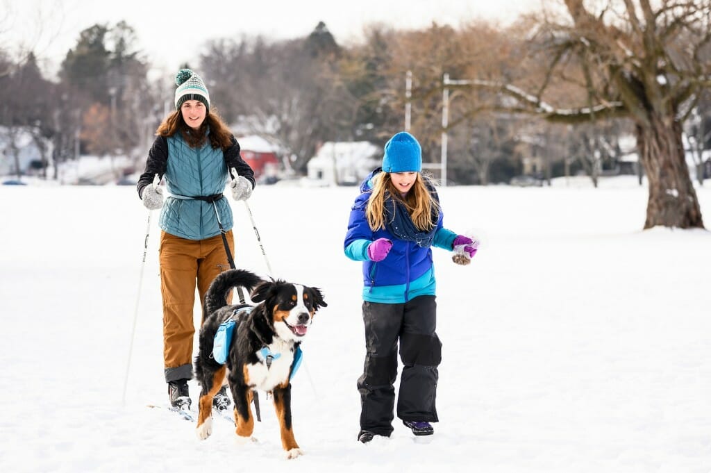 Two woman walk through the snow with a dog.