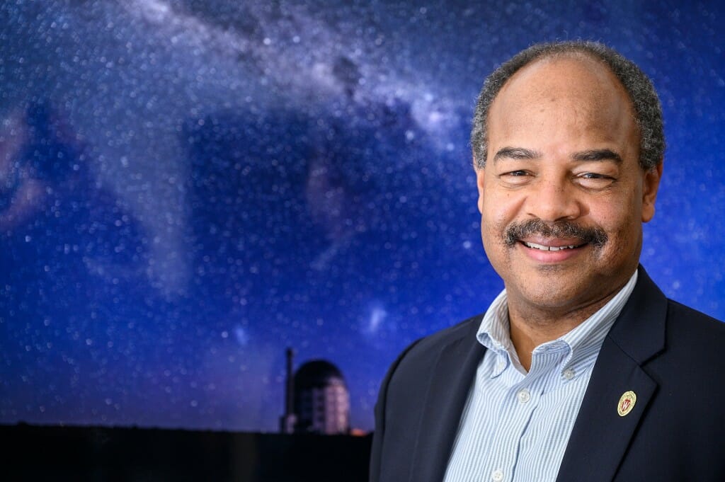 Portrait of Eric Wilcots standing in front of a picture of an observatory and the night sky