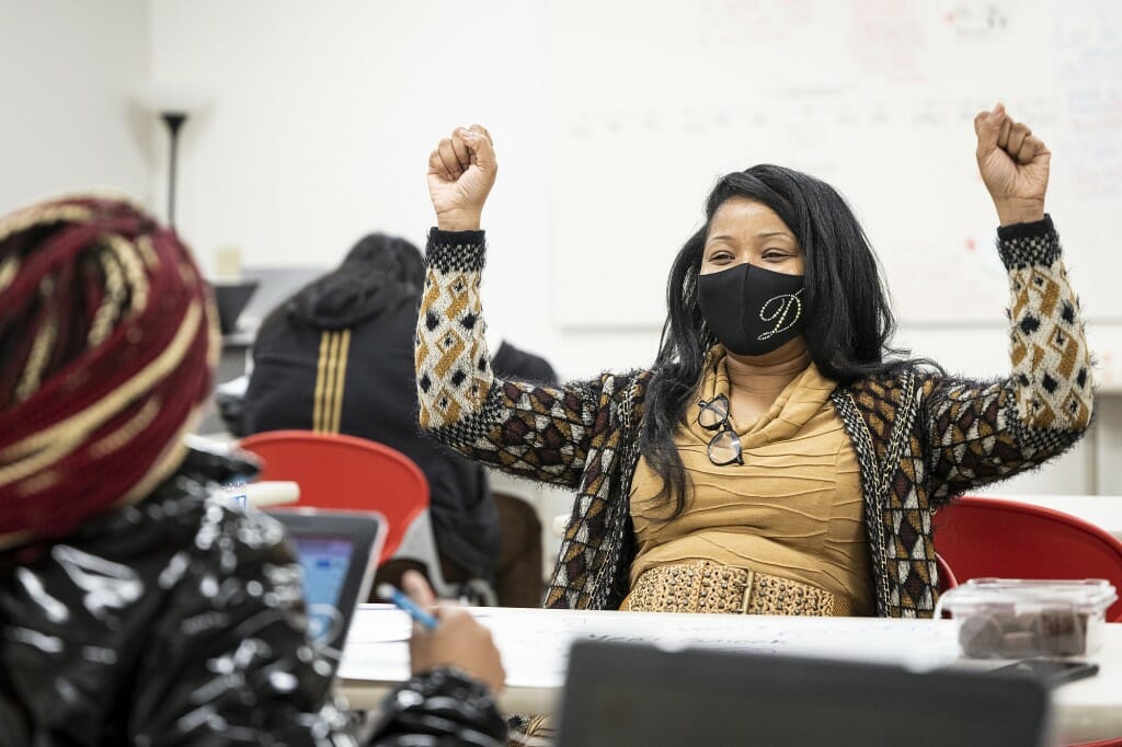 A teacher wearing a face covering raising her arms while sitting across the table from a student