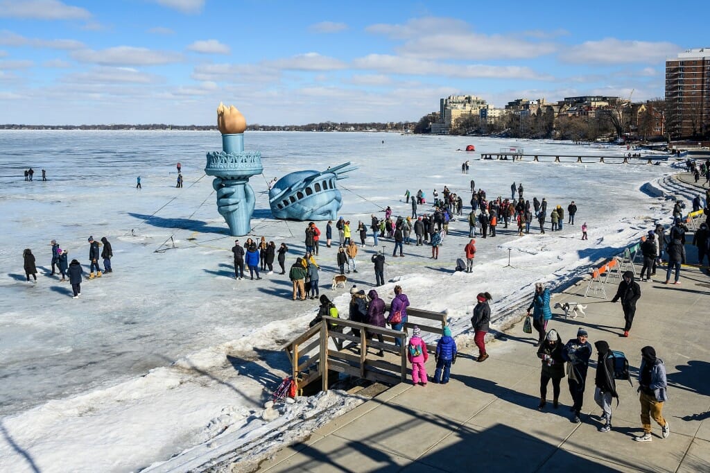 Winter Carnival-goers mingle with an inflatable replica of the Statue of Liberty on frozen and snow-covered Lake Mendota near the shoreline.