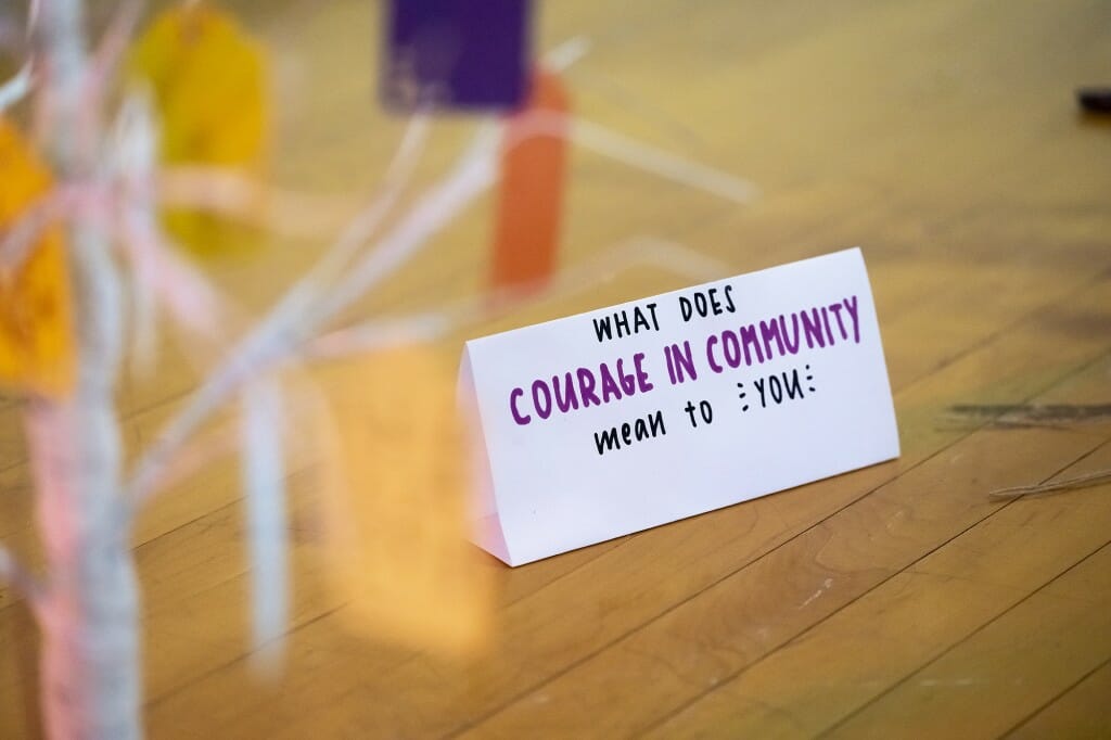A sign with the question “What does courage in community mean to you?” prompts viewer participation.