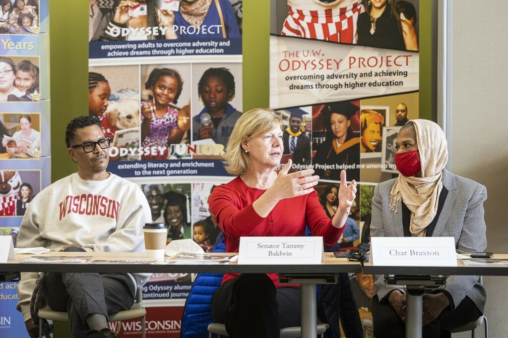 Tammy Baldwin sitting at a table between 2 people in front of a collage of images from the Odyssey Project