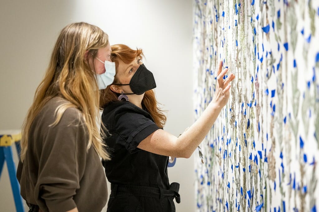 Closeup of 2 people looking at embroidered plants on a wall