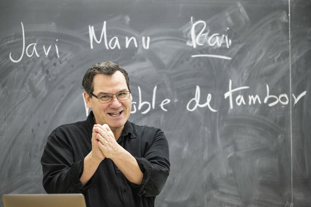 Juan Egea in a classroom, clasping his hands while standing at a lectern in front of a blackboard