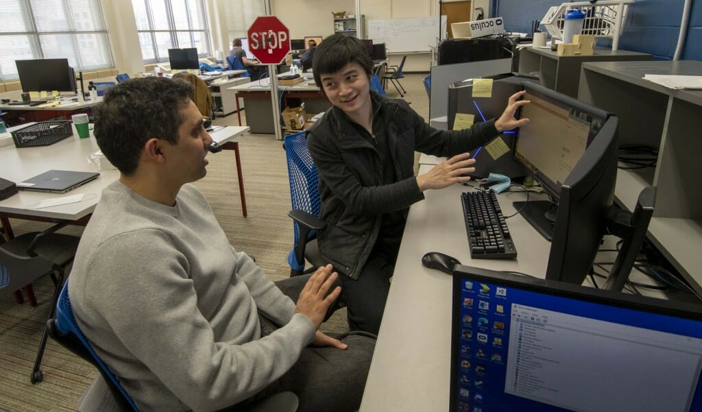 Two people sitting a a desk with one motioning toward a computer monitor