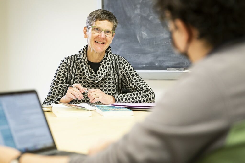 Judith Houck smiling and sitting at a table in front of a chalkboard across from an unidentified student