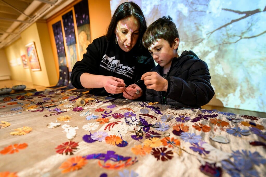 At left, Bliss Thiel, a microbiologist in the UW–Madison Veterinary Diagnostic Lab, helps her son Henry (age 6) sew flowers with written messages into a collaborative tapestry called “Marking Time
