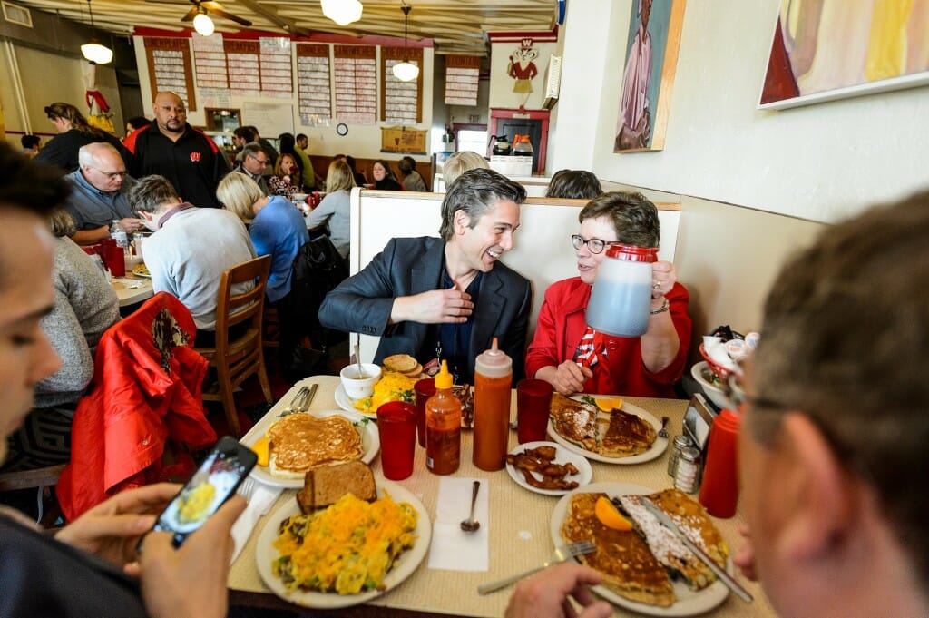Blank and Muir sitting in a diner booth with food on the table, Blank holding a large pitcher while Muir smiles