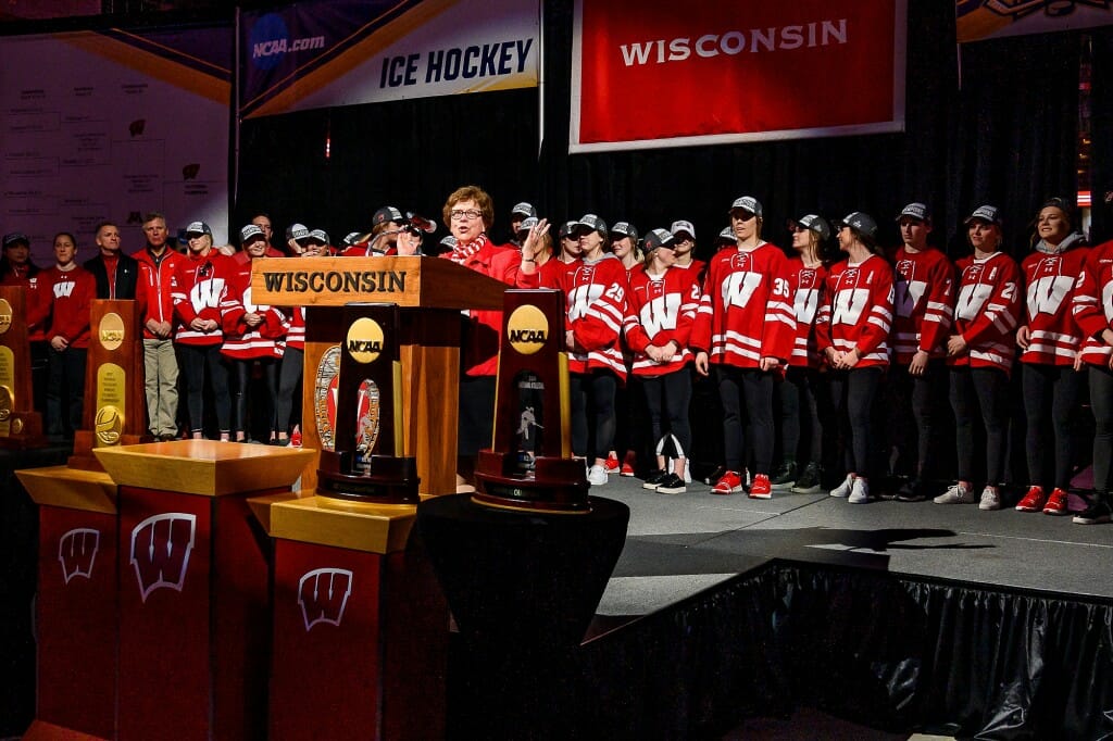 Blank speaking from a podium with people in hockey jerseys standing behind her on stage