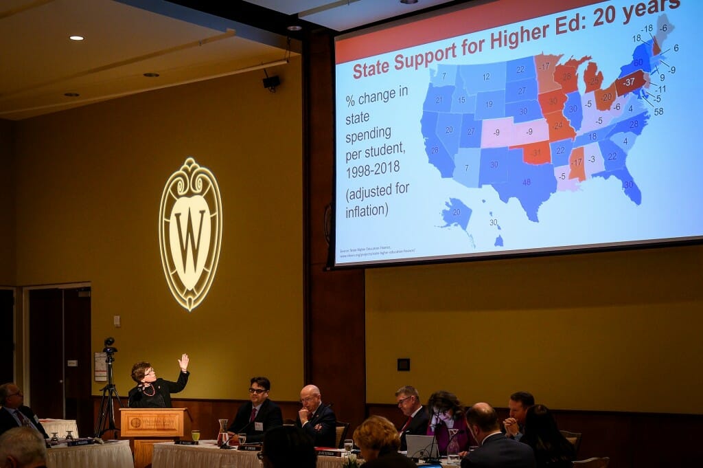 Blank speaking at a podium under a projected map of the U.S. showing which states increased higher education spending (blue) and decreased it (red). Wisconsin is red.