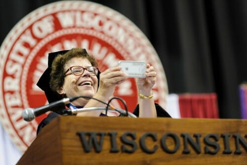 Blank at podium in academic hood in front of UW–Madison seal, aiming a cellphone at audience for a picture