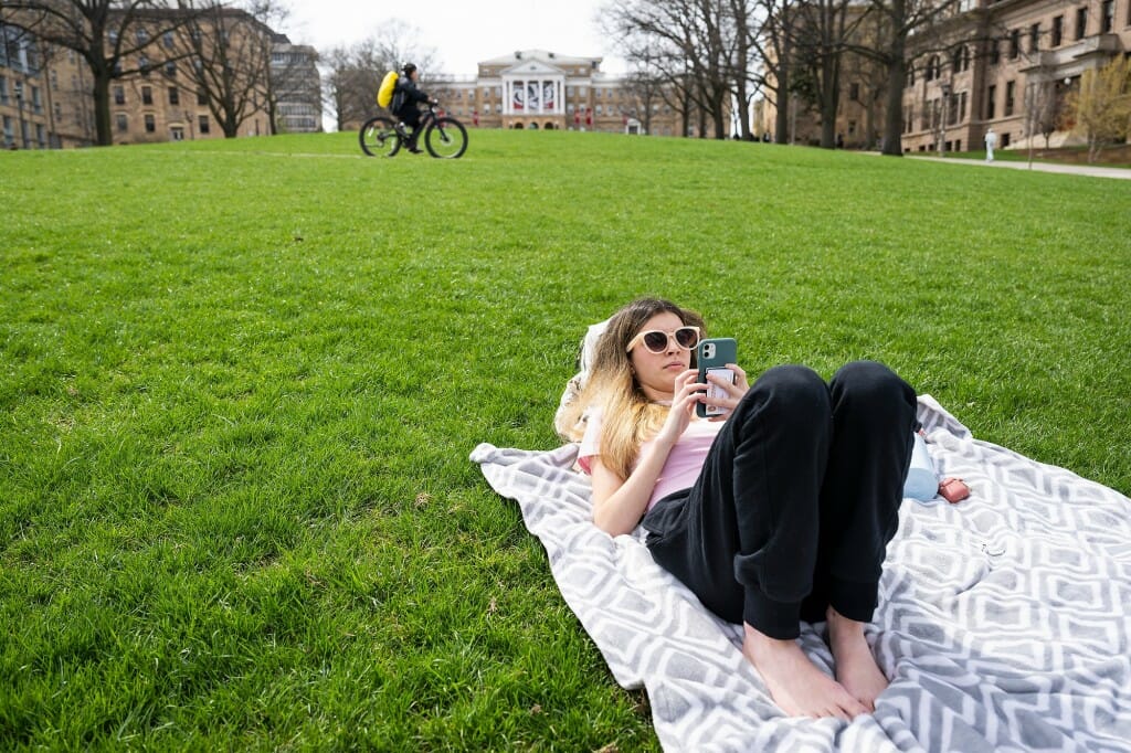 A person wearing sunglasses reclining on her back on a blanket while looking at her phone