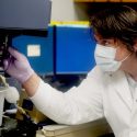 Brock Kingstad-Bakke wearing a surgical grade mask and white lab coat and purple rubber gloves, looking at a lab experiment