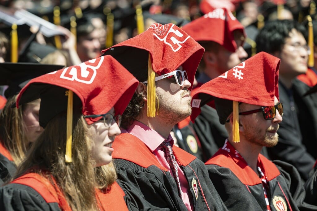 Several people sitting in the sunshine with red towels draped over their graduation caps