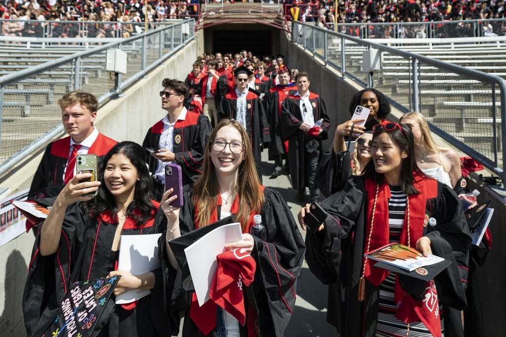 A group of students in graduation gowns smiling and emerging from a tunnel enter the stadium