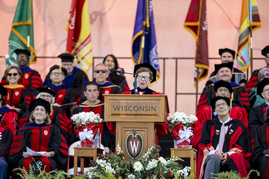 Rebecca Blank smiling while speaking at a podium with a WISCONSIN sign and "W" crest