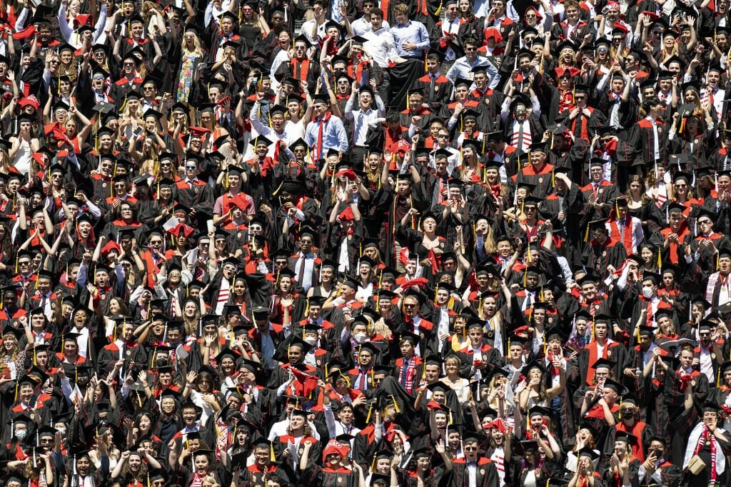 A wide shot of a large group of graduates in caps and gowns standing inside the stadium