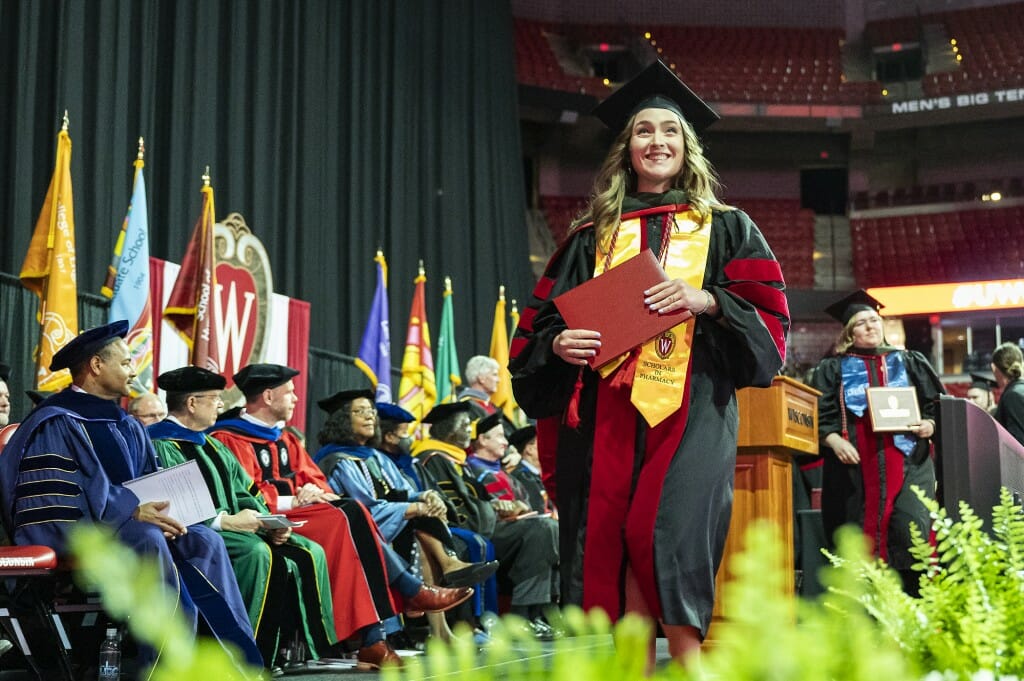 Person in cap and gown walking across stage while holding red diploma cover