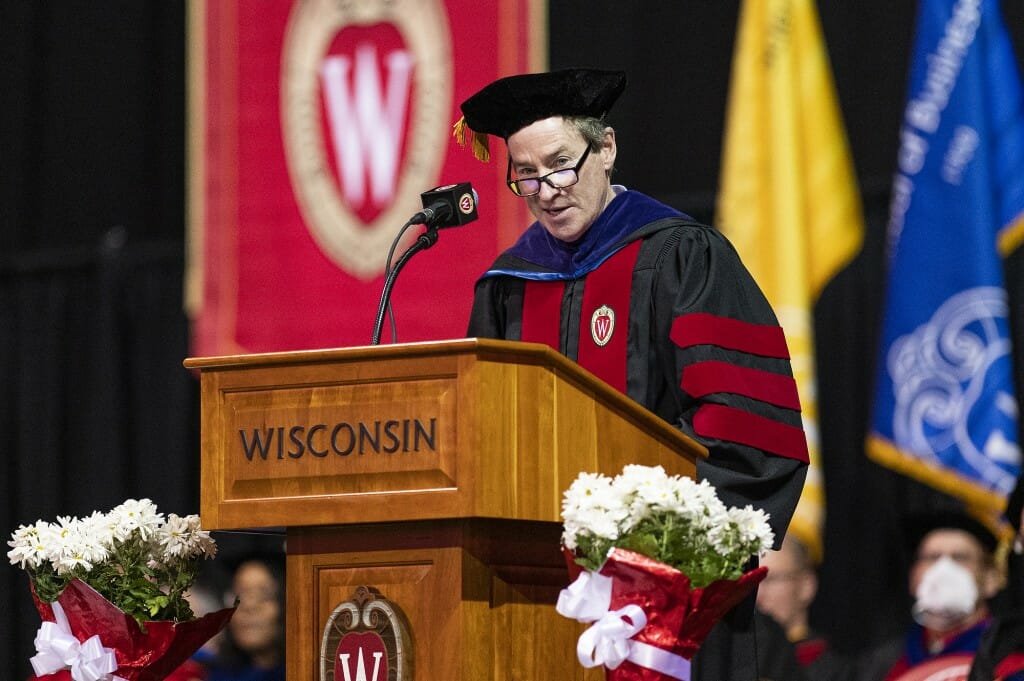Jones, wearing academic hood and gown, speaks at podium in front of a red 