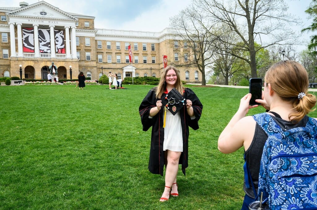 Soon-to-be-graduate Makenna Ley poses on Bascom Hill with her gown and decorated neurobiology and biochemistry motarboard hat while Liza Spellman take photos.