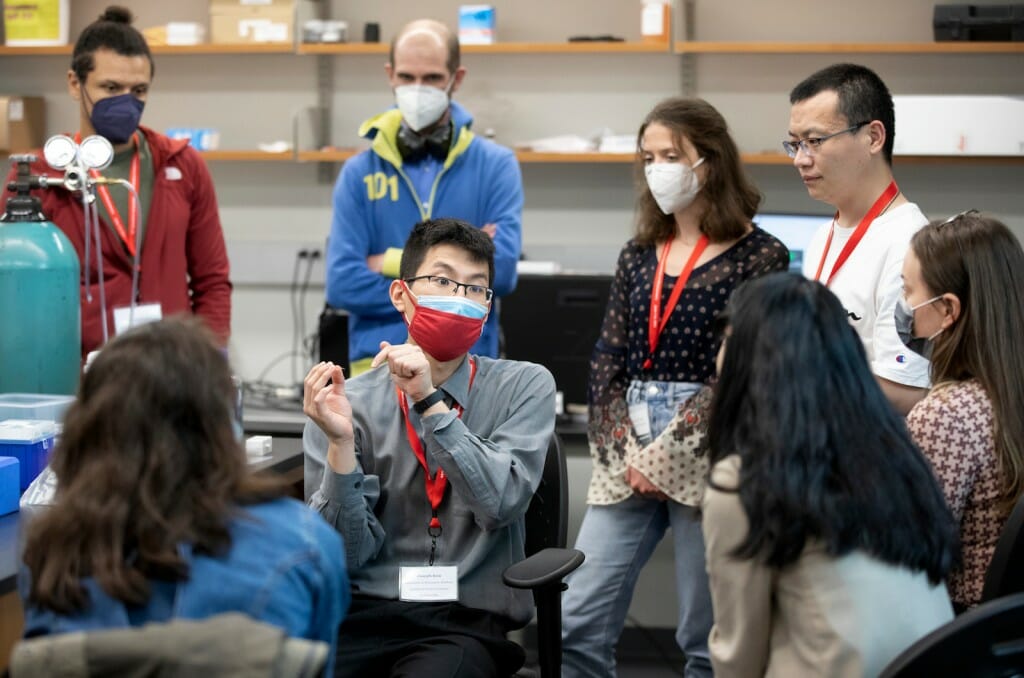 Graduate student Joseph Kim sits in the middle of a circle of scientists in a science lab, leading an interactive workshop.