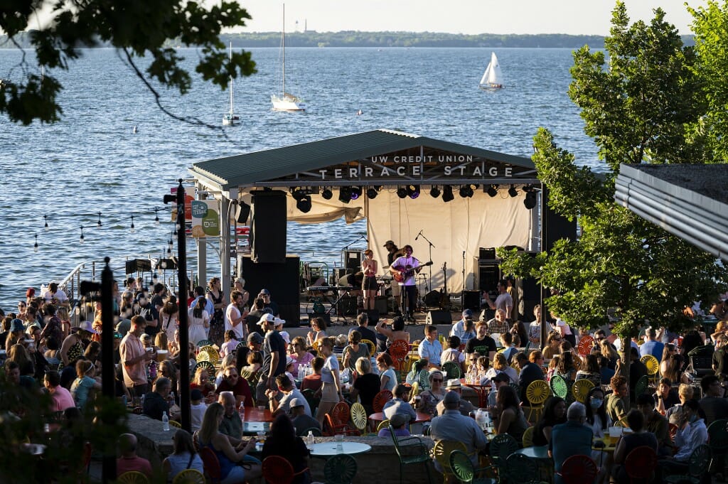 Large group of people listening to a band performing under a canopy with Lake Mendota in the background