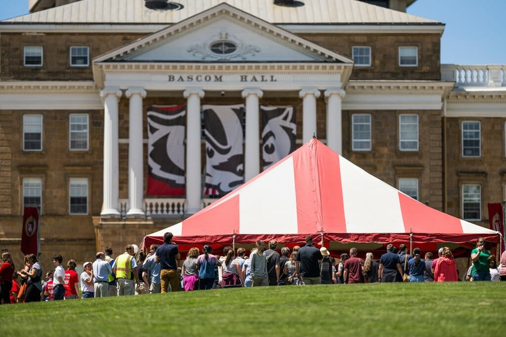 People gathering under a red and white tent in front of Bascom Hall