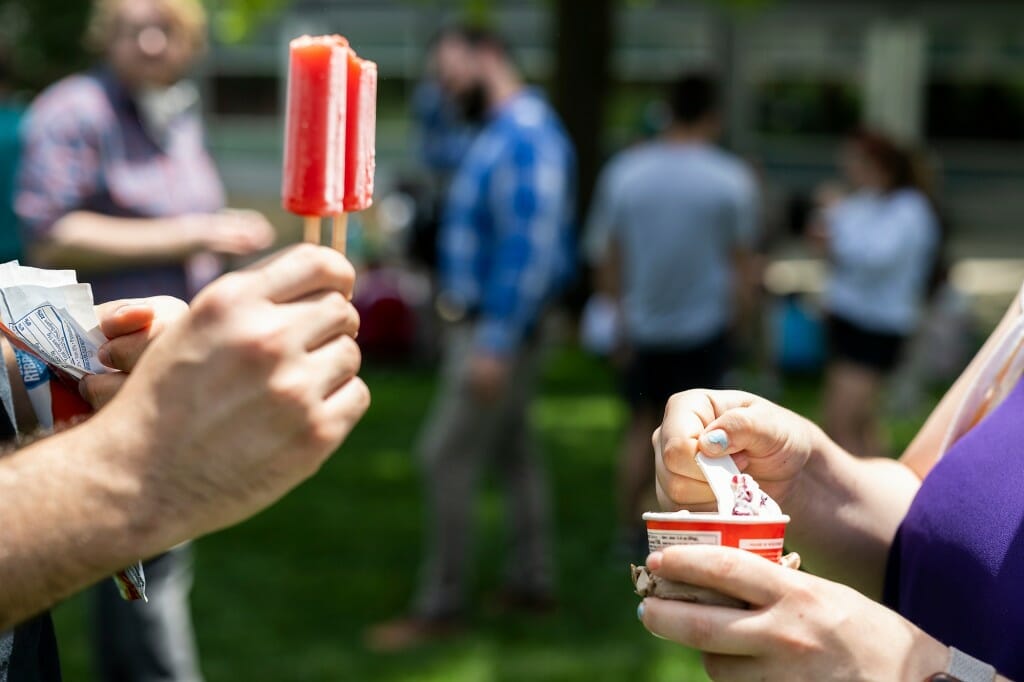 Closeup of one hand holding a popsicle and another holding a cup of ice cream and a spoon