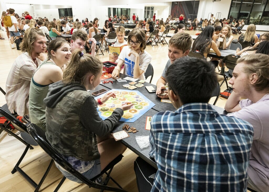 A student rolls the dice above a game board, as others look on and smile.