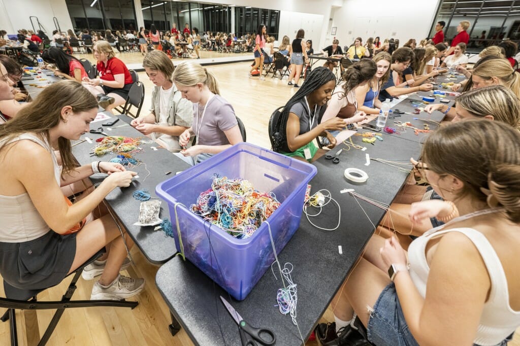 Students lined up at several tables work at making bracelets with yarn.