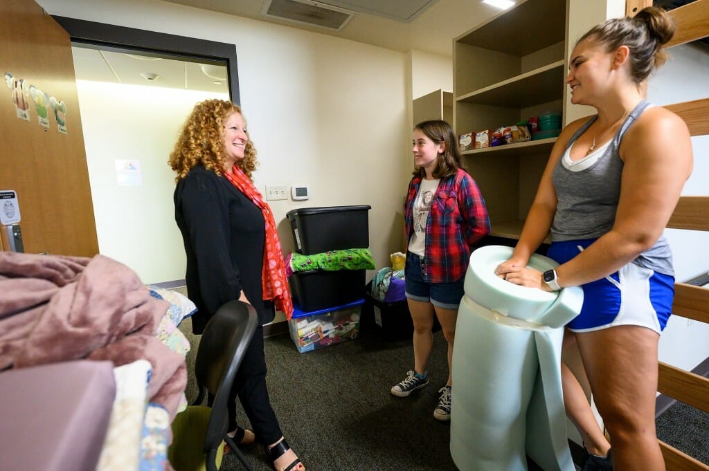 A woman talks to two other young women in a dorm room filled with unpacked items.