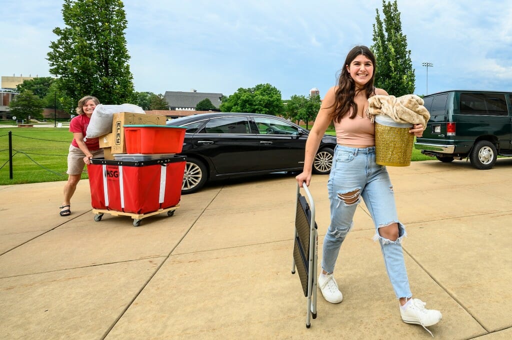 A smiling student in the foreground carrying items through a paved area, behind her is a woman pushing a red loading cart.