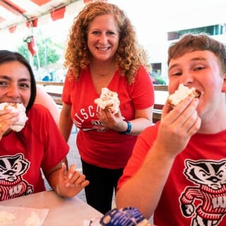 Chancellor Mnookin wears a red UW-Madison shirt and poses next to a young man and young woman while eating cream puffs
