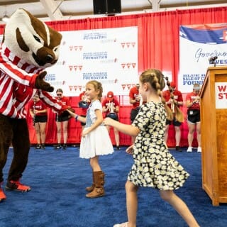 Bucky Badger wears a red and white strip shirt while dancing with two young girls wearing dresses on a stage
