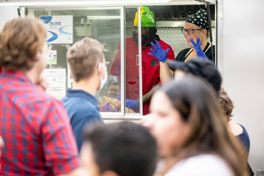A crowd stands outside a food truck, with a person taking orders inside.