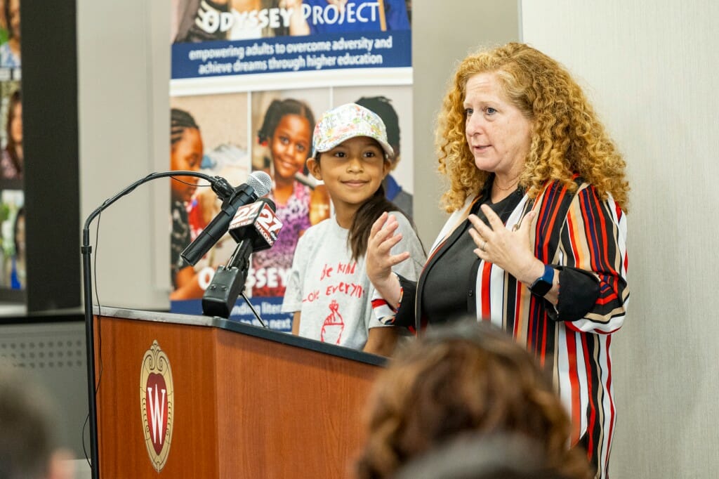 The chancellor speaks at a podium with a girl standing next to her, looking at her.