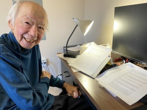 an elderly smiling man sits in profile at a desk with sheets of paper, a computer and a lamp
