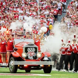 An old fashioned fire engine vehicle blowing out smoke and carrying cheerleaders drives down the field.