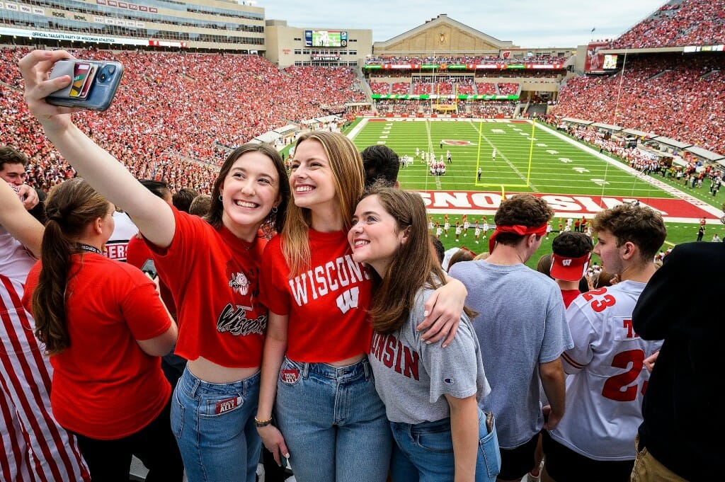 Three young woman stand with their arms around each other as one of them takes a selfie with a cell phone.