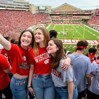 Three young woman stand with their arms around each other as one of them takes a selfie with a cell phone.