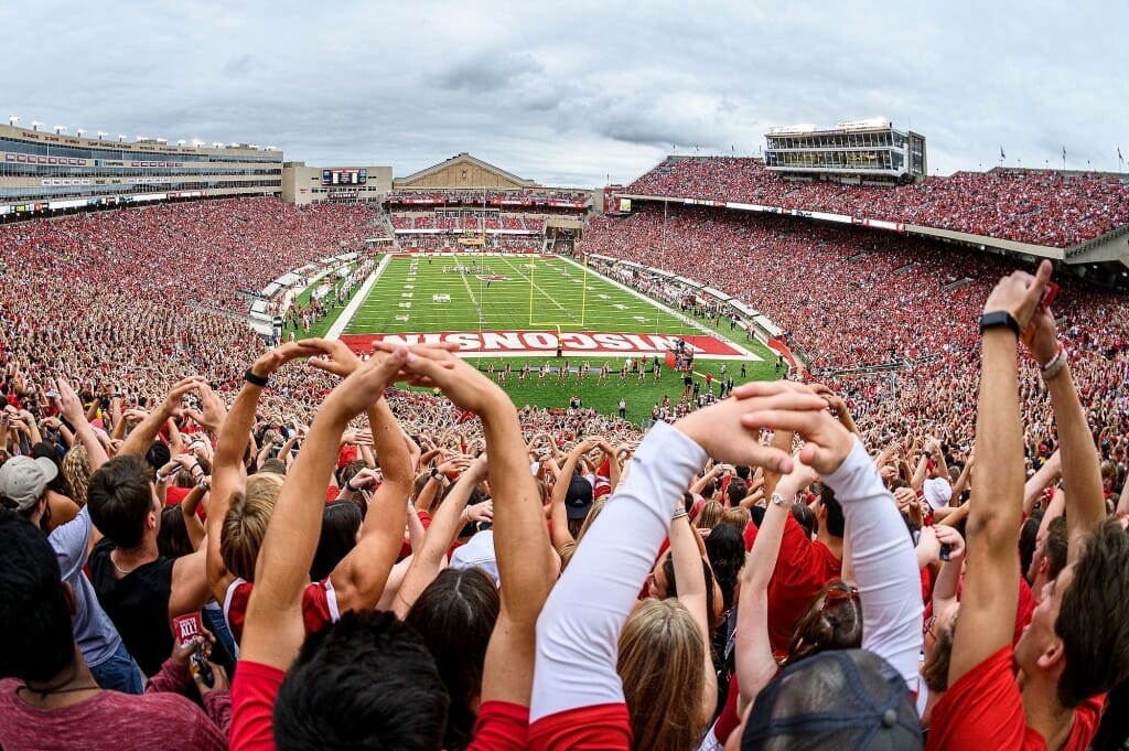 Fans hold their arms up to make an "O" with the football field in the background.